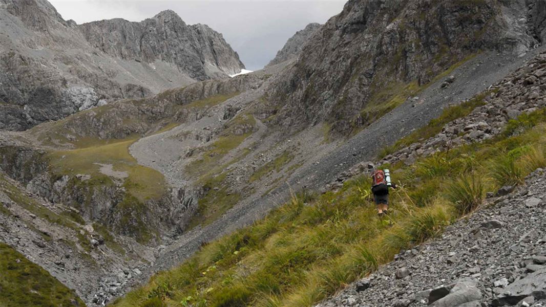 A tramper walks along a steep scraggly mountain side. You can see that as the tramper continues it gets steeper and steeper until the end of the valley connects together from the two ridges of mountains. It’s mostly rock.