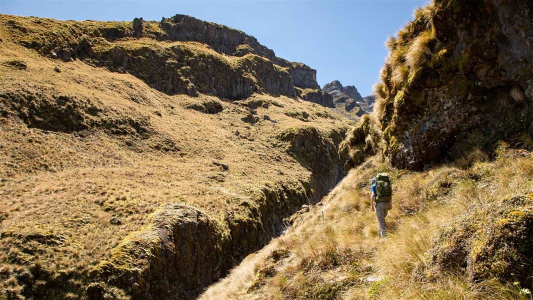 A lone tramper follows white markers towards a big crack in the earth. There's a ridge line either side of the crack.