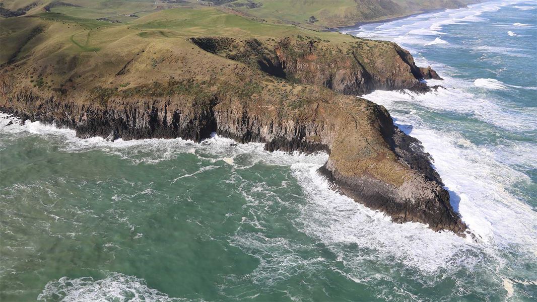 A large piece of land juts out from the rocky coast. The piece of land jutting out has almost vertical rockfaces that extend down into the sea below.