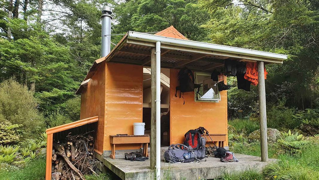 A small orange hut surrounded by forest, tramping gear hanging from the roof over the deck.