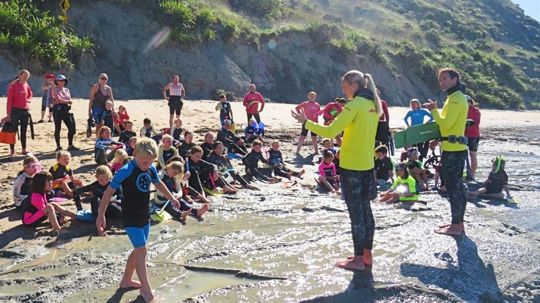 A large group of people and children listening to two coordinators at the water's edge on the beach. 