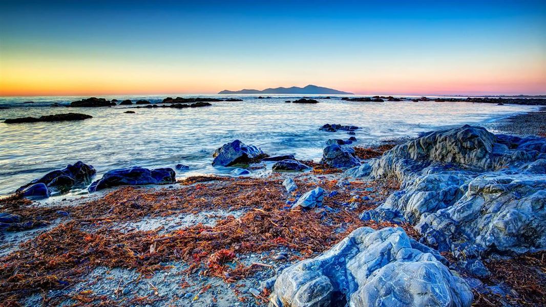 A photo taken from a shore covered in red seaweed and smooth boulders. An island is just visible across the water under an orange sky from sunset.