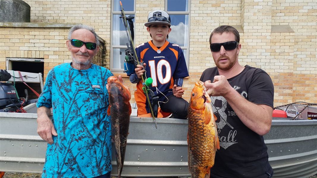 Three men stand in front of a boat holding up two large koi and a hunting bow.