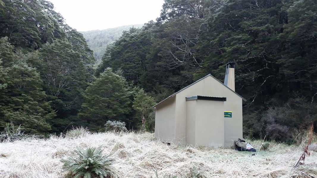 Branch Creek Hut sits amongst the grasses with a tramping bag resting by the door.