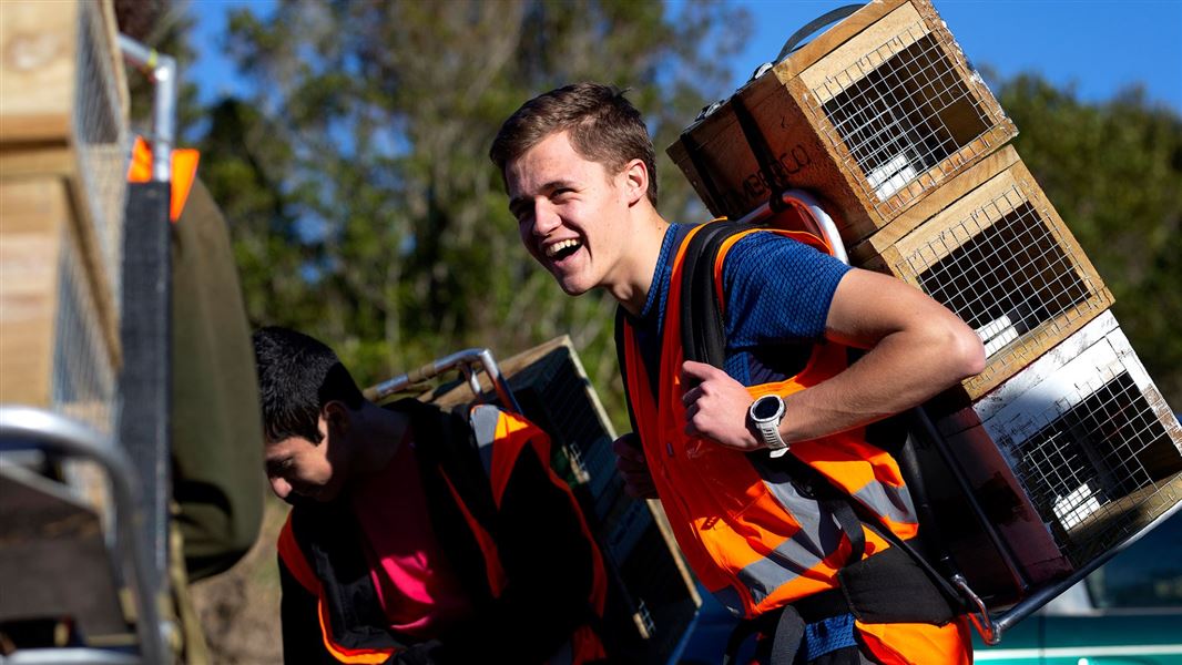 A DOC ranger seen from the back, speaking to two male students carrying traps and dressed in high visibility jackets 