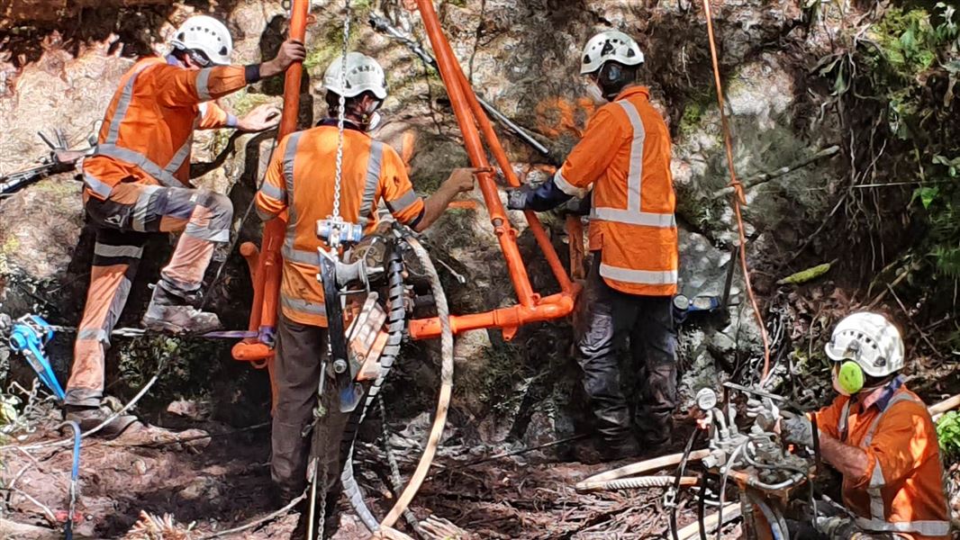 Four construction workers lowering a large metal rock anchor into the gorge.