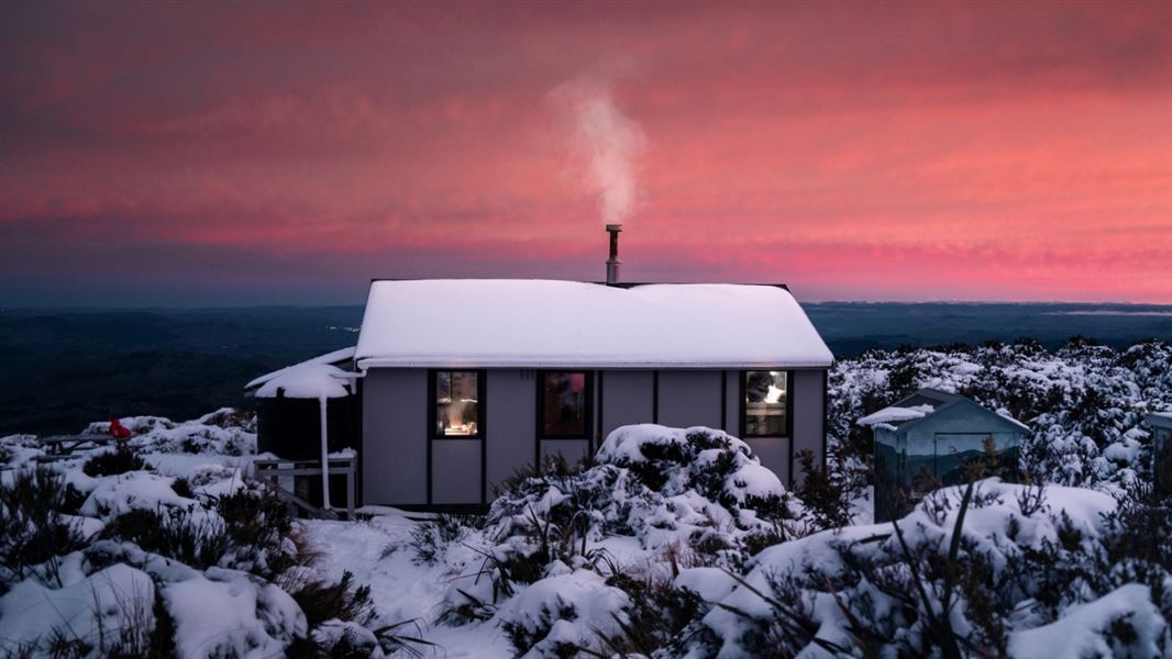 A grey hut sitting amongst the snow and frost with smoke rising from the chimney into a bright pink sky at dusk. 