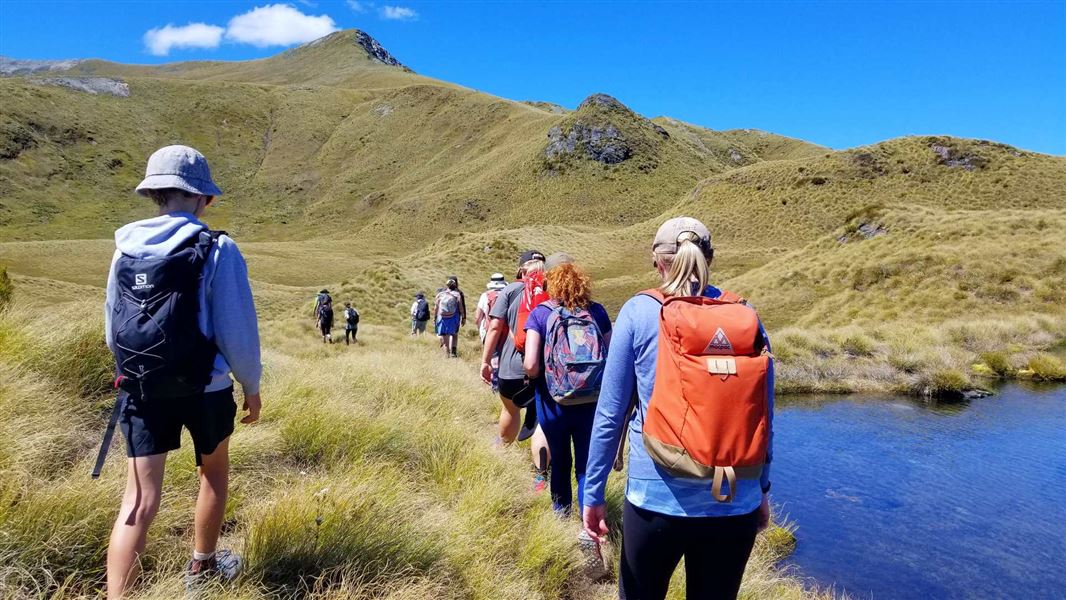 A long line of people hiking along a track surrounded by brush by a lake.