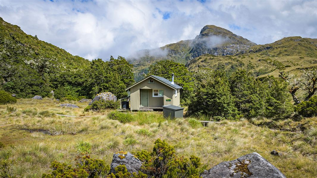 Small building in grassy area by hill and trees.