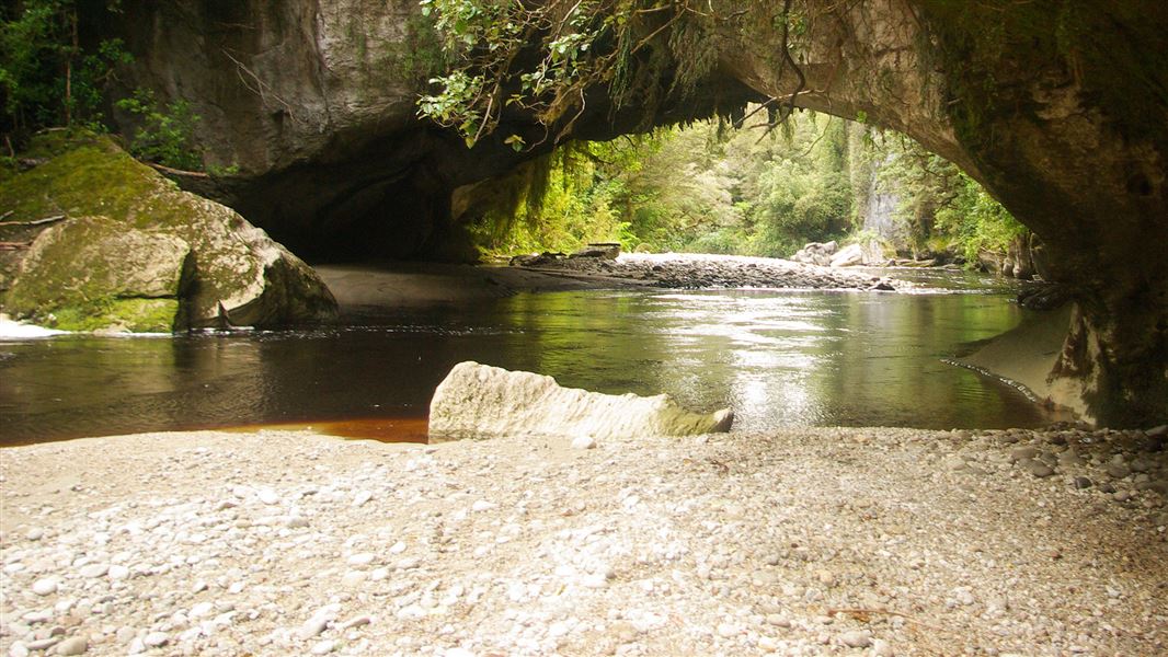 A photo of a riverbank by a large natural archway framing a waterfall.