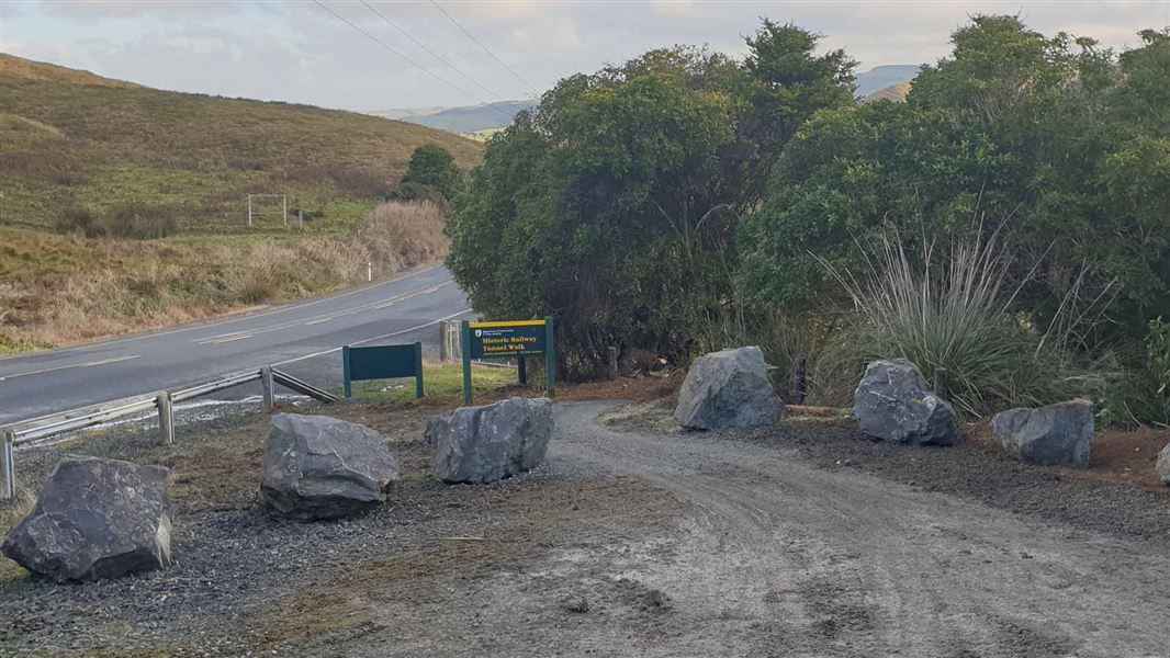 A gravel carpark leads to a DOC sign with Historic Railway Tunnel Walk amongst the bush.