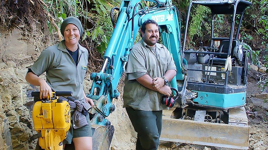 A woman and a man carrying large, heavy drilling equipment with a large blue digger behind them. 