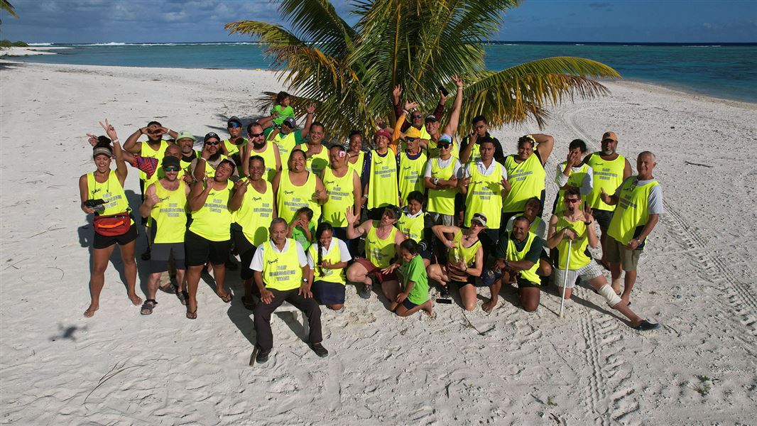 The Palmerston Atoll rat eradication team stand on a sunny  Cook Island's beach with golden sand. The team are in bright high vis yellow t-shirts and are waving at the camera.