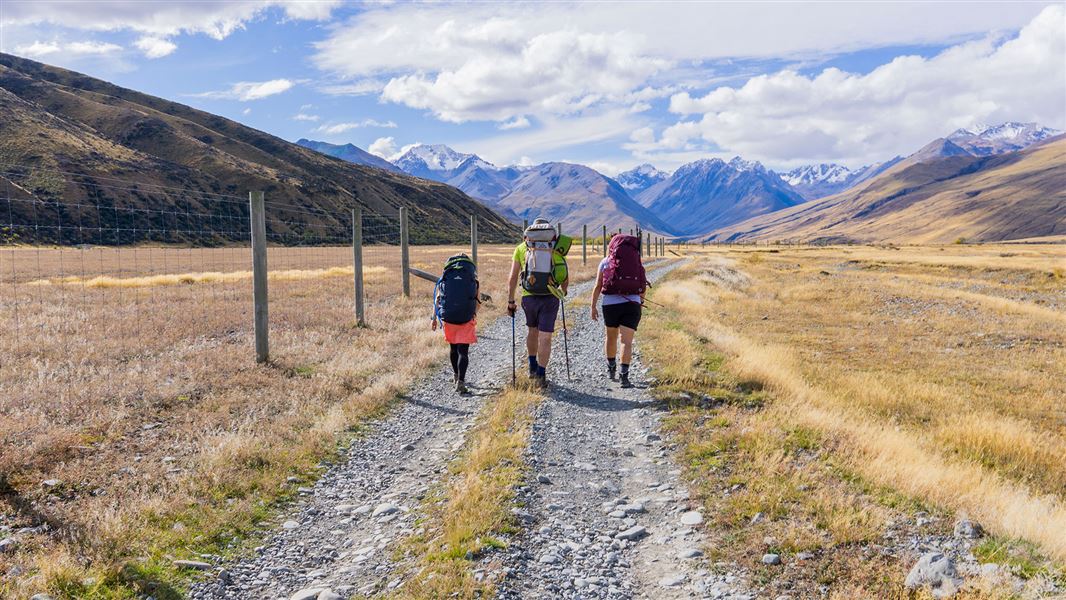 Three people walking on gravel track near hills.