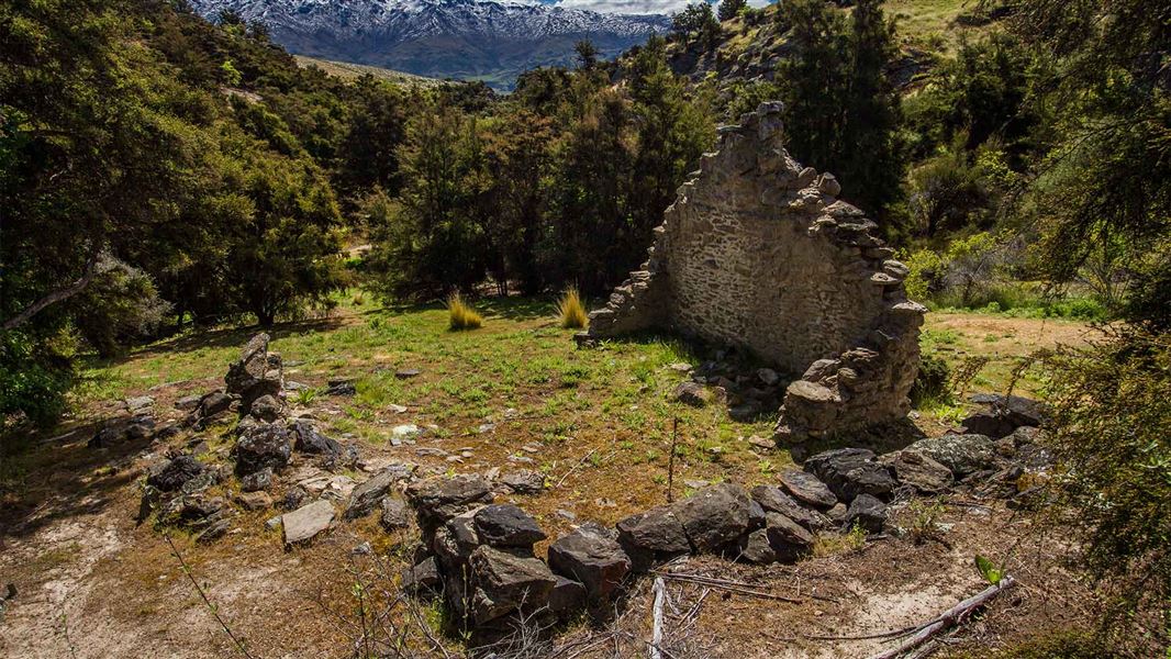 Only one wall of an old stone house remains standing in a small clearing. Off in the distance there is a range of mountains covered by snow.