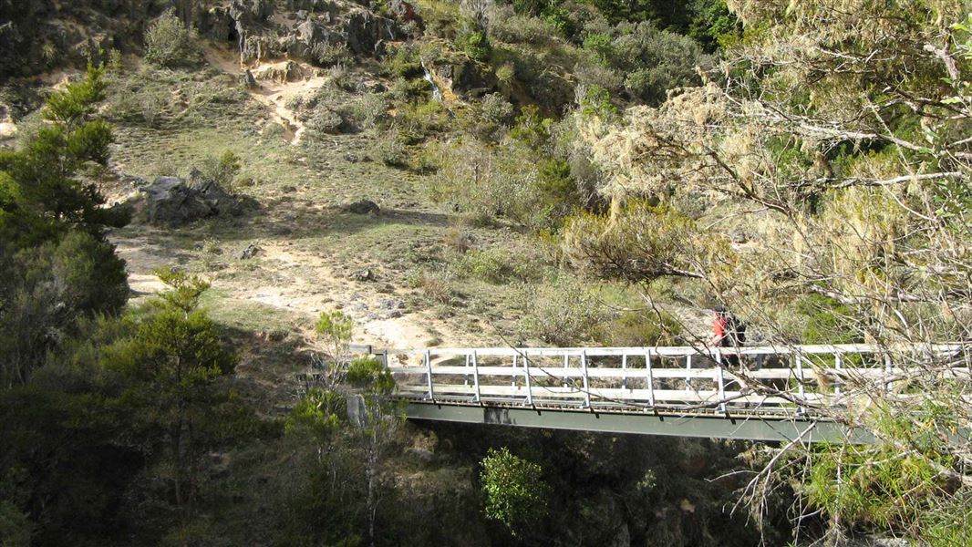 A bridge with white painted wooden sides crosses a river below a steep, eroding hill side.