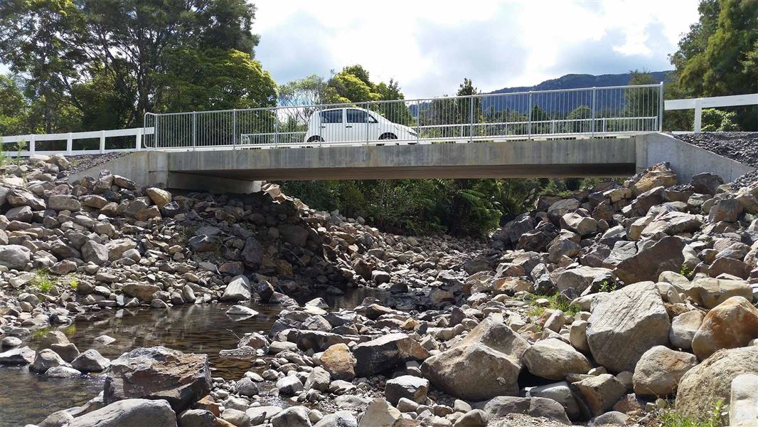 A rocky stream dips below a concrete bridge that a car is driving over.