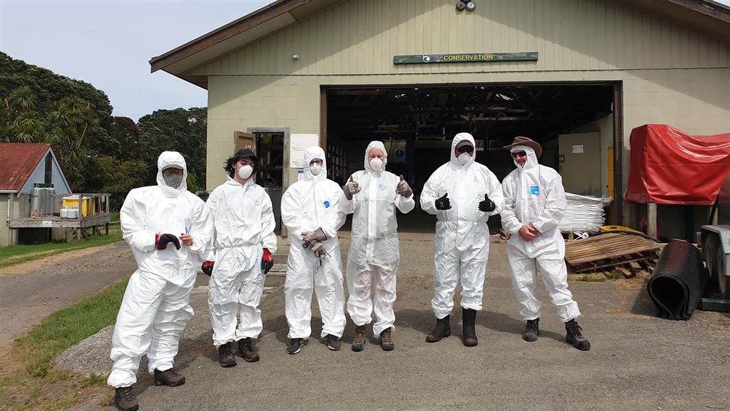 Volunteers in white overalls stand in front of a shed on Motutapu Island.