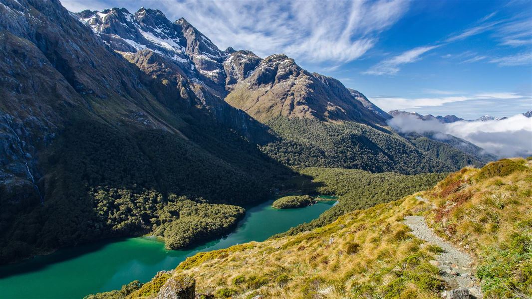 A view of a large mountain with a deep blue lake below from the top of a winding, grassy  path.