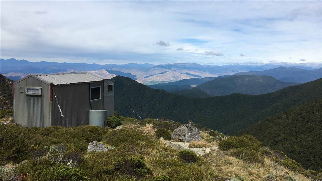 A corrugated iron small hut sits on the ridge of a high mountain. From here you can see out over many mountain ranges in the distance.