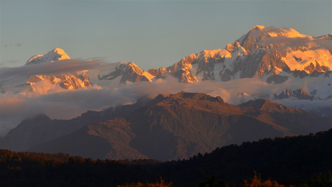 Snow capped mountains glow golden in the late afternoon sun behind dark forested foothills.