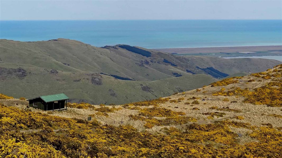 View down to a shelter. The shelter is on the a gentle rise in the surrounding hills. The sea can be seen far in the distance.