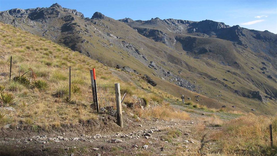 View of a rocky track that extends lazily alongside a hill that disappears toward rough scraggly taller hills in the distance.