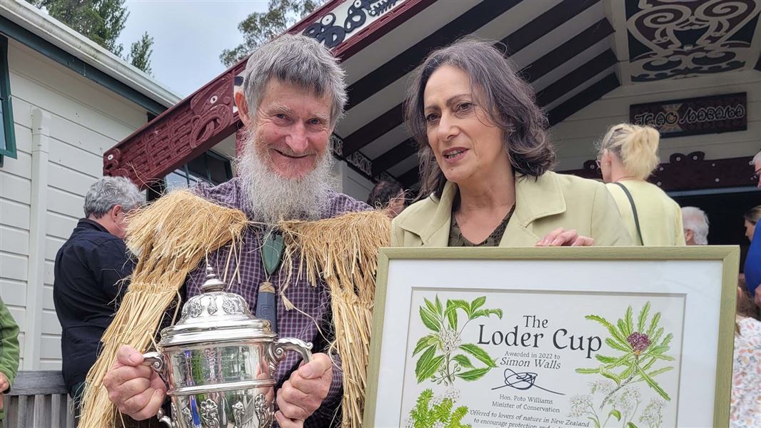 A man holds a trophy next to a woman with a certificate that says The Loder Cup was awarded to Simon Walls. 