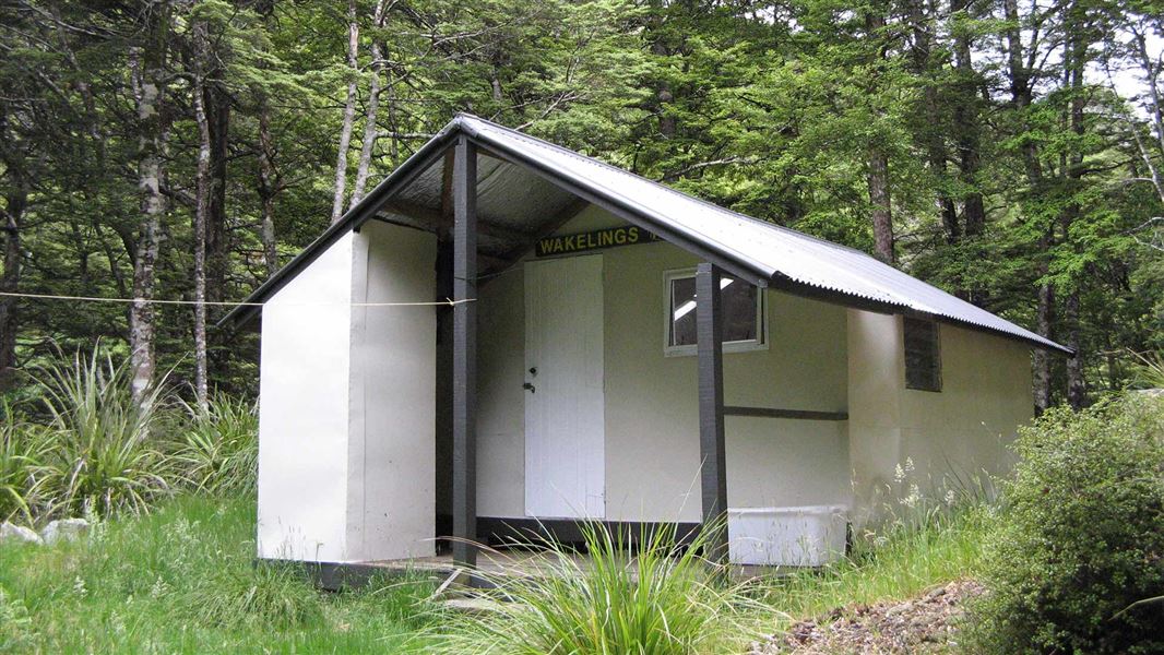 A light grey hut with a tin roof stands in a grassy clearing.