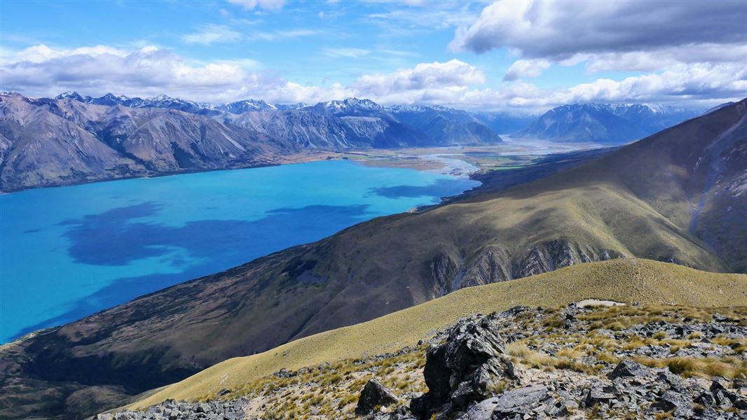 View from top of a alpine ridge across a turquoise lake with mountain range in the background.