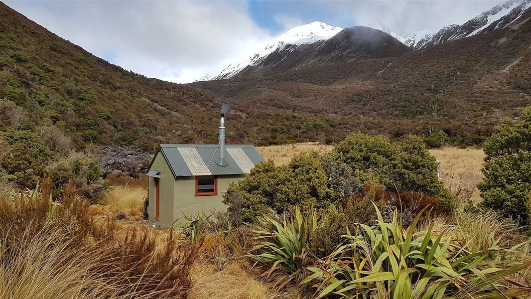 Small building among grasses with snowy hill in background.