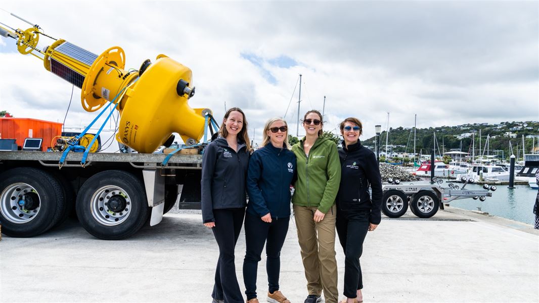 Four technical advisors from DOC, GWRC and NIWA stand with the new buoy on a trailer by the water.