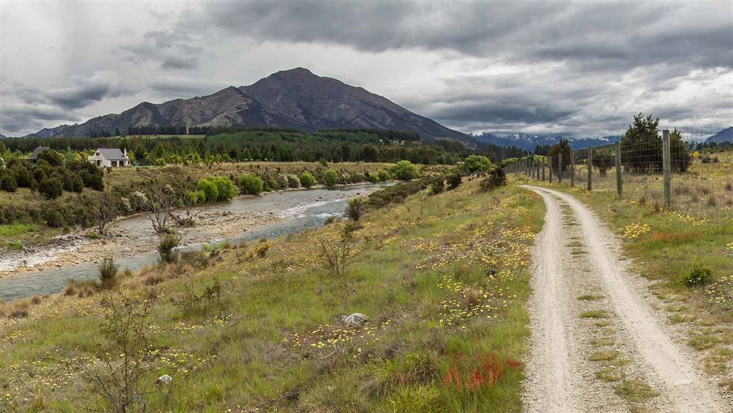 A gravelly track windws alongside a shallow river. A large hill can be seen in the distance with snow peaked mountains further away.