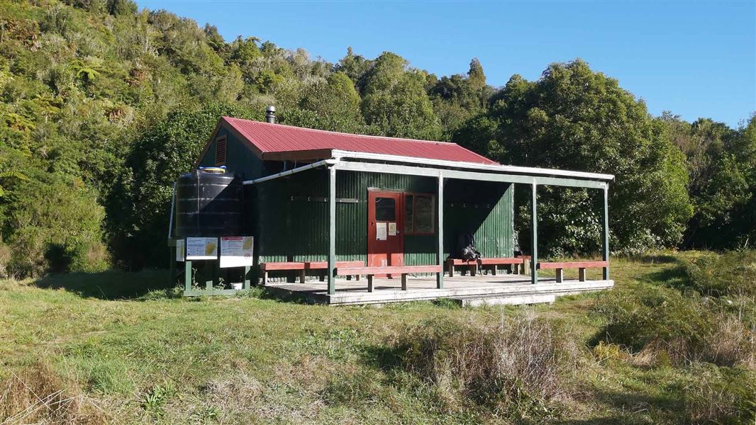 Green and reed lodge in grassy clearing with trees behind. 