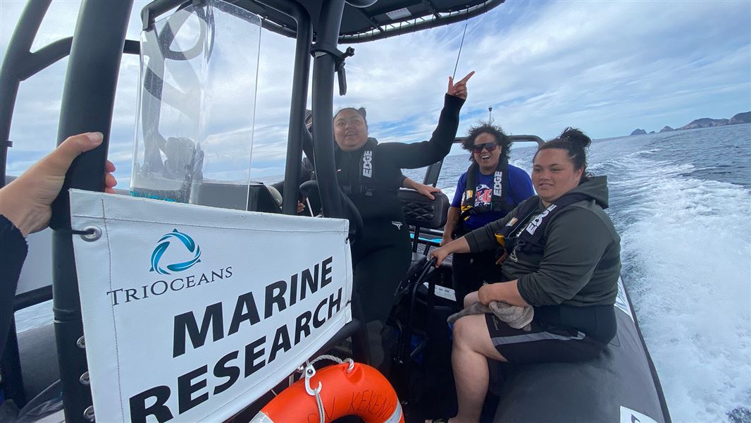 Three people in life jackets sit at the back of a speed boat with islands in the background.