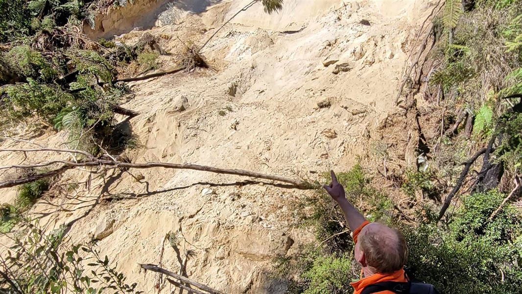 A man in high vis points to an eroded bank with fallen trees.