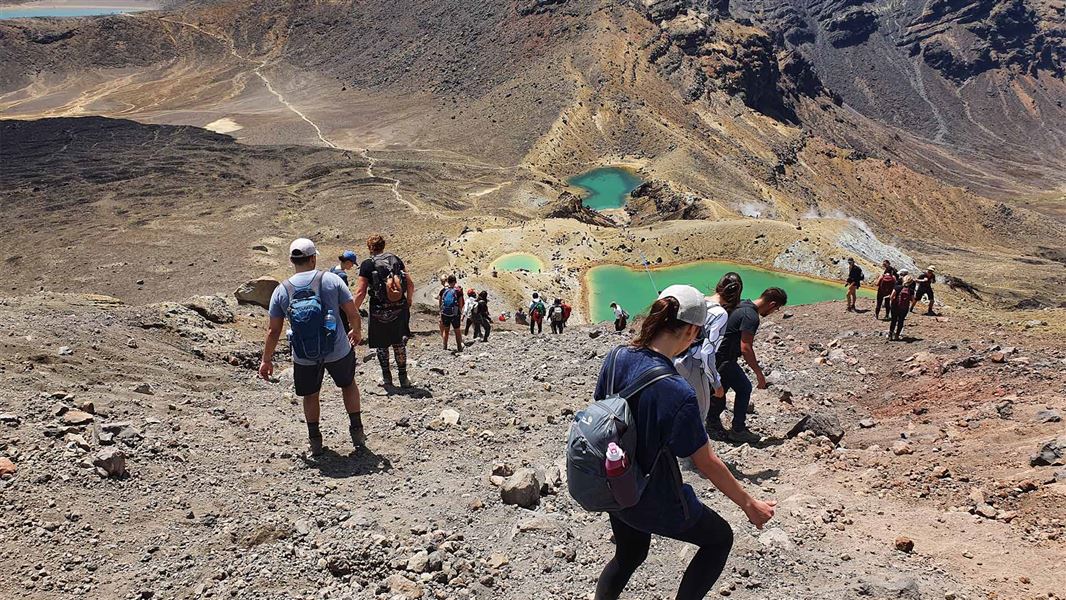 A large number of people slowly descend the side of a rocky mountain. They are heading towards green coloured lakes.